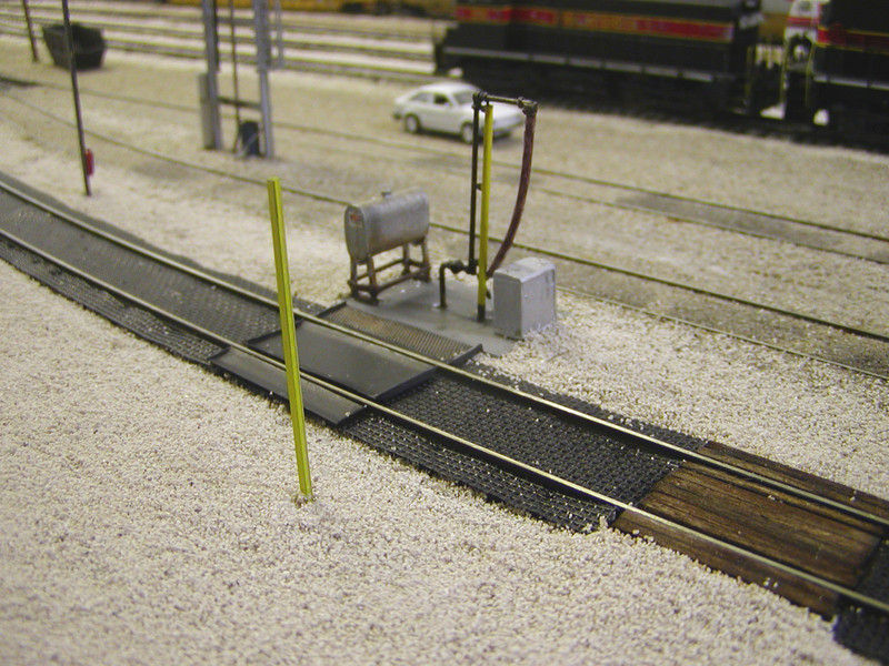 New fuel pad, fuel spill collection trays and mats, support/guard posts made from old rail, grade crossings for MOW hostlers and their equipment, and buried enginehouse tracks.  The fuel pipe was heavily kitbashed from a pair of American Limited Models fuel cranes, while the fuel spill collection trays are scratchbuilt from 0.010" styrene and Scale Scenics mesh.  The fuel spill mats are garden weed cloth.

The yellow post in the foreground, and the grey one in the upper left with the fire extinguisher, are sections of code 55 rail to match the prototype's use of rail for these purposes here.

The oil spills on the two adjacent enginehouse tracks are India ink wash applied with a dropper.