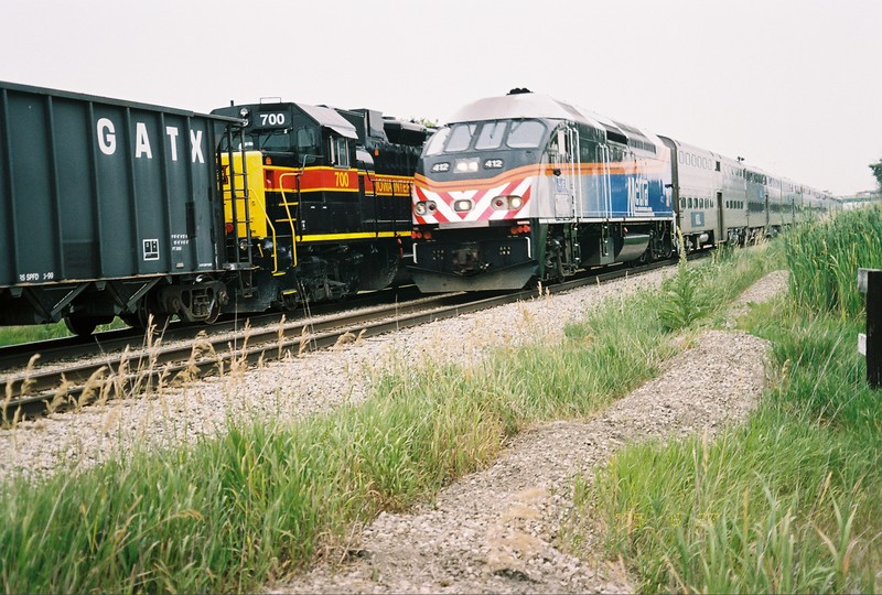 A westbound Metra slows for its Hickory Creek stop, while the IAIS east train switches Ozinga.  June 28, 2005.