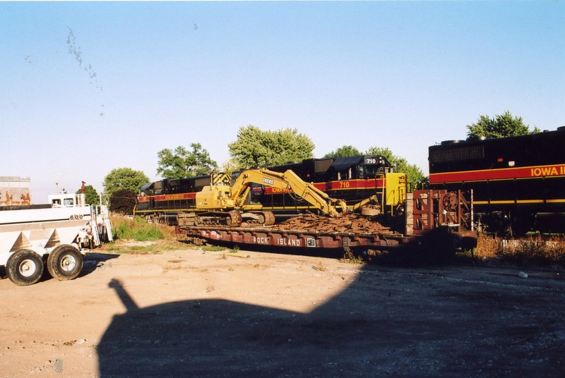 The eastbound passes the ex RI flat in Atalissa with the electromagnet trackhoe and a pile of tie plates on board.  Aug. 7, 2005